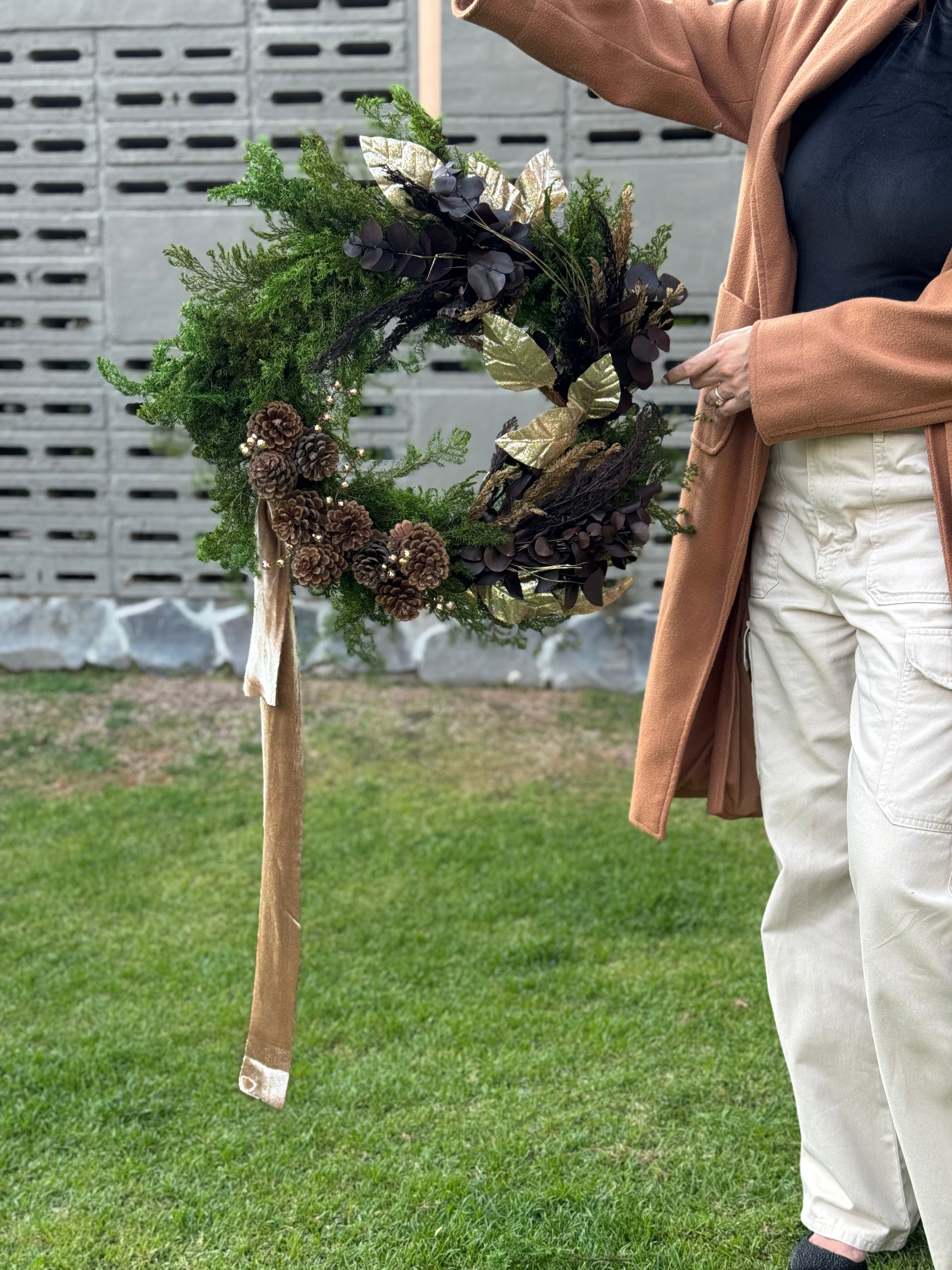 Corona navideña de 60 cm con pino preservado verde, eucalipto café, piñas naturales y detalles dorados y bronce.