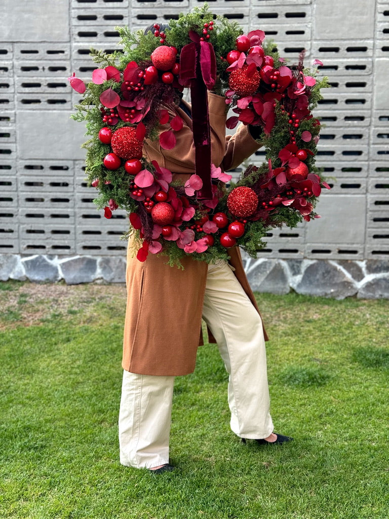 Corona navideña grande de 90 cm hecha de pino preservado con eucalipto tinto, esferas rojas brillantes y listón de terciopelo tinto.