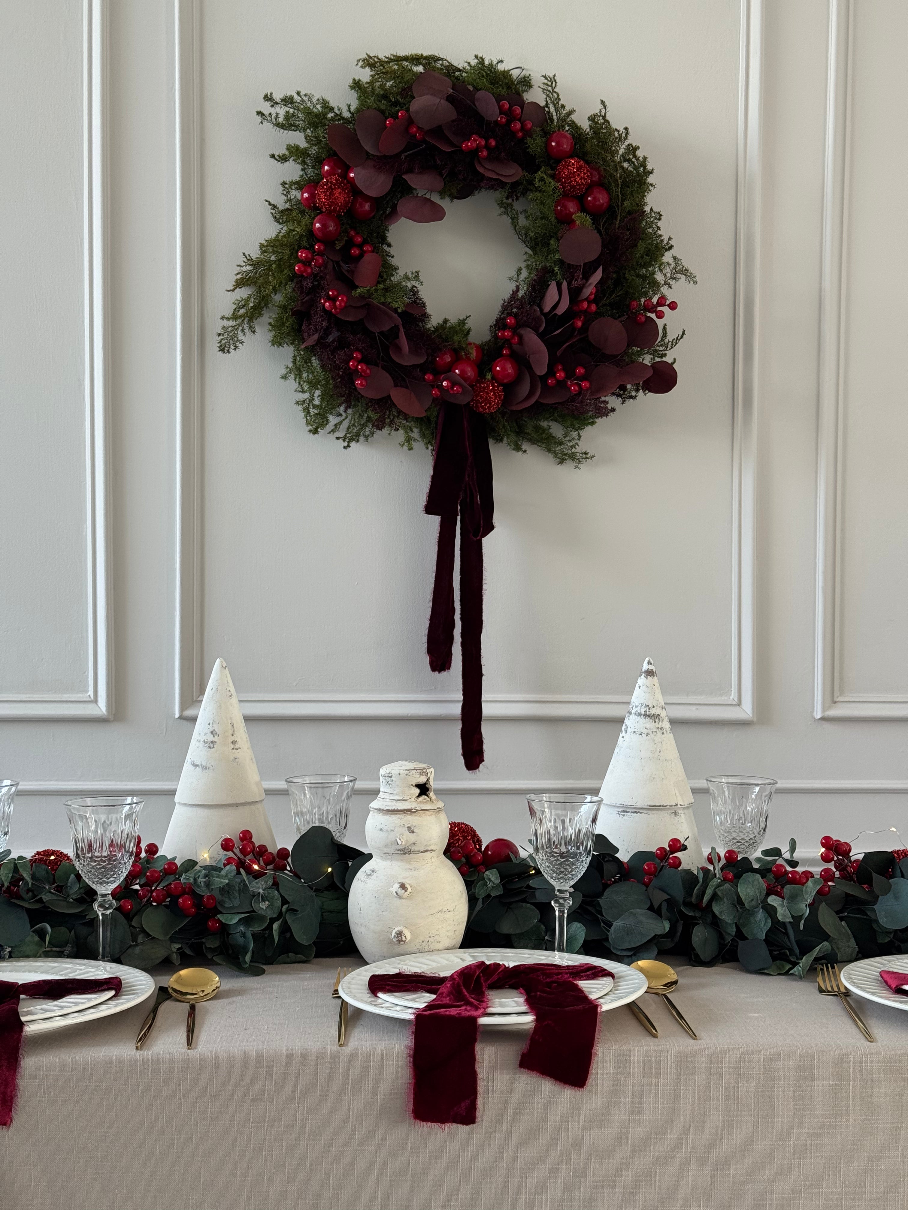 Corona navideña de 60 cm con pino preservado, eucalipto tinto, ornamentos rojos adiamantados, cerezas decorativas y listón de seda tinto.
